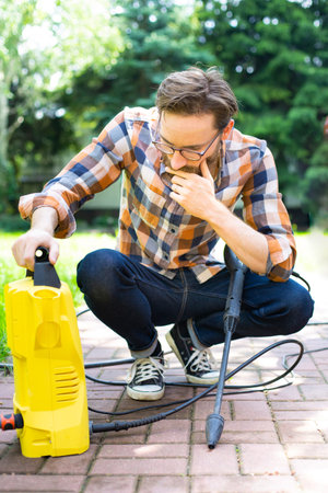 Young man in casual clothes using a yellow compact home high pressure washer. Washing in the garden...