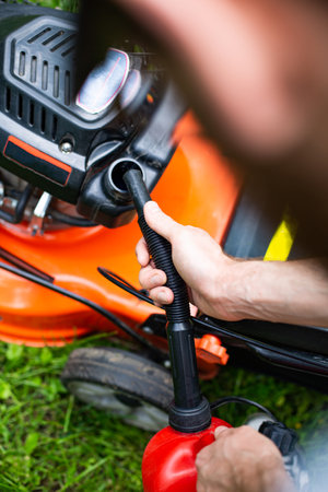 Man refilling a fuel tank in a petrol lawn mower with a red plastic canister with a funnel, close up on a hand with a funnel. Gardening, using a gasoline lawn mower in a garden on a sunny dayの写真素材
