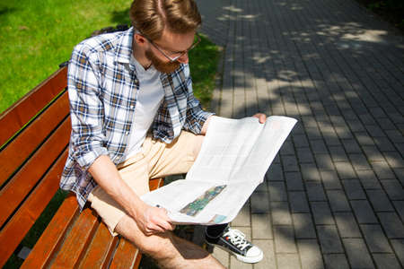 Young man reading a newspaper on a bench in the park on a sunny day.の写真素材