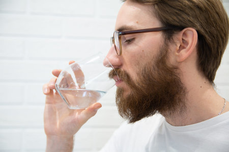 Portrait of a young handsome adult man in a white t-shirt, drinking a glass of pure water, side view. A healthy life concept, modern interior in a background.の写真素材