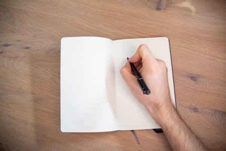 Top view of a male hand with a black pen on a white paper notebook on a natural wooden desk. A writer, journalist, student preparing for writing, horizontal view with a copyspace.の写真素材