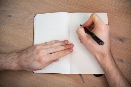 Horizontal view of male hand with a black pen on a white paper notebook on a natural wooden desk. A writer, journalist, student preparing for writing, close-up with a copyspace.の写真素材