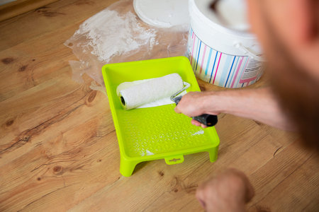 A paint roller with white paint in a house painter's hand, on a green paint tray, on a wooden floor....