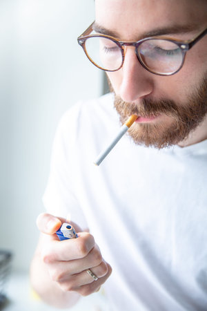 Nicotine addiction. Young adult, bearded man in a white t-shirt lighting a cigarette with a blue...