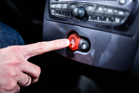 Male hand pressing a hazard warning lights button with a red triangle on a car dashboard. A road...