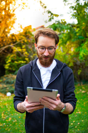 Young adult, bearded man using a tablet outdoor, in a garden, on a sunny day. Using an electronic, mobile, portable device to browse the internet, to read books, newspaper.の写真素材