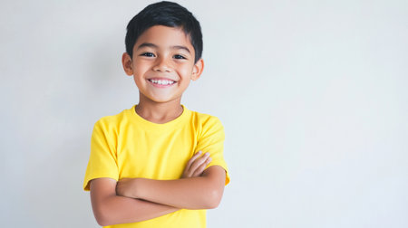 a cute young boy smiles brightly with his arms folded wearing a yellow shirt in front of a plain wallの素材