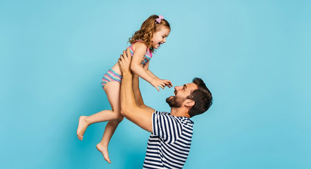 A joyful dad lifts his laughing daughter into the air, sharing a playful moment on a bright blue backdrop. Celebrating fatherhood and joy.の素材