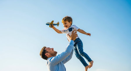 A father joyfully lifts his son in the air, celebrating a moment of happiness together under a bright blue sky. The boy holds a toy airplane.の素材