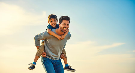 A joyful father carrying his son on his back, both smiling against a blue sky. A perfect representation of family bonding and happiness.の素材