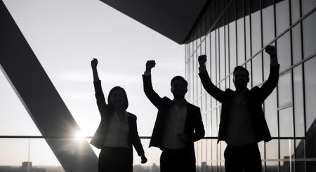 A silhouetted group celebrates success with arms raised high against a bright sky, symbolizing achievement and teamwork.の素材