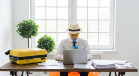 A man wearing a fedora hat and tie is working on a laptop at a desk, with a yellow suitcase beside him, suggesting a work-travel scenario.の素材