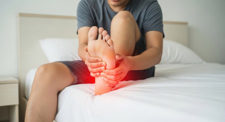 Close-up shot of a man sitting on a bed, holding his foot, with a red glow indicating pain and discomfort.の素材