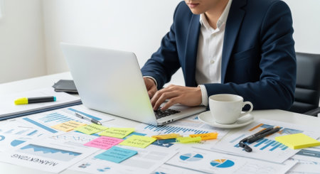 A focused businessman in a suit is analyzing data and working on a laptop at his office desk, surrounded by charts and graphs.の素材