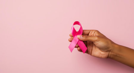 A hand holds a pink ribbon, the symbol of breast cancer awareness, against a soft pink backdrop, promoting support and hope.の素材
