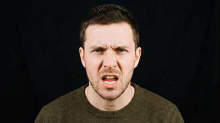 a close up shot of a man with an angry expression. he is wearing a green sweater and is set against a solid black background. he looks upset.の素材