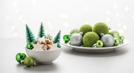 A festive arrangement of Christmas decorations featuring green and silver spherical ornaments, miniature green Christmas trees, and small star-shaped cookies in a white bowl, set against a clean white background.の素材