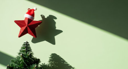A red star ornament wearing a Santa hat casts a shadow on a light green wall, with a small Christmas tree in the foreground.の素材