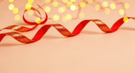 A close-up shot of a red ribbon with a golden pattern, elegantly curled and flowing across a soft peach background. Out-of-focus golden bokeh lights create a festive and magical atmosphere.の素材