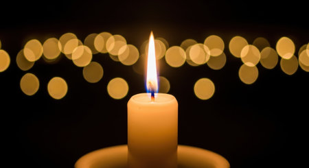 A close-up shot of a lit candle, its flame casting a warm glow against a dark background filled with soft, out-of-focus lights.の素材