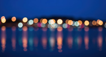 Abstract image of blurred city lights reflecting on water, creating a bokeh effect with vibrant colors against a dark blue sky.の素材