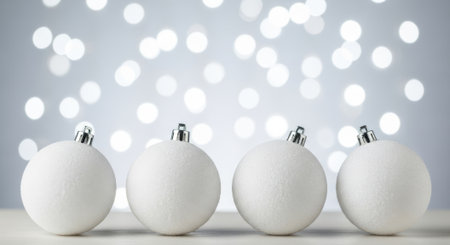 Four pristine white Christmas ornaments are arranged in a row against a soft, out-of-focus background of sparkling bokeh lights, creating a festive and elegant holiday scene.の素材