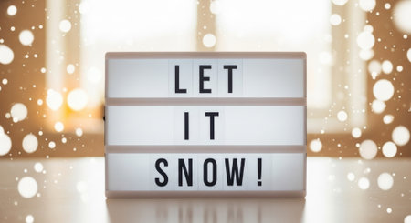 A lightbox sign with the text Let It Snow is displayed against a blurred background of warm lights and falling snowflakes, evoking a cozy winter atmosphere.の素材