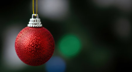 A close-up shot of a single red Christmas ornament, sparkling with glitter, hanging delicately. The background features soft, blurred bokeh lights, creating a festive and magical atmosphere.の素材