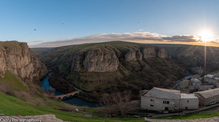 A breathtaking panoramic shot captures a historic stone bridge spanning a winding river at the bottom of a dramatic, deep canyon. The setting sun casts a warm glow over the rugged landscape, highlighting the ancient architecture and natural beauty of the scene. A small building is visible on the right.の素材