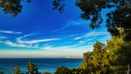A serene seascape with a vibrant blue sky, delicate clouds, and lush green trees framing the view.の素材