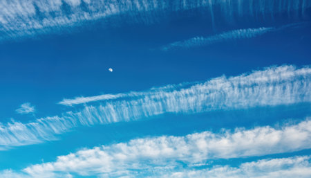 A breathtaking view of a clear blue sky filled with delicate, wispy clouds stretching across the horizon. A small, bright moon is visible in the distance.の素材