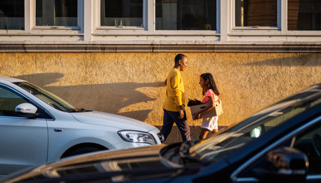 A father and daughter walk side by side on a sidewalk, with cars parked nearby.の素材