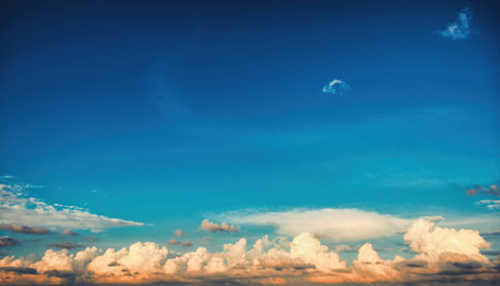 A breathtaking view of a clear blue sky filled with soft, white clouds illuminated by the warm hues of a setting sun.の素材