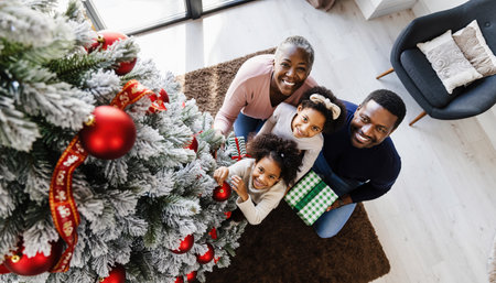 A joyful African American family, including parents and two daughters, smiles warmly while gathered around a beautifully decorated Christmas tree, holding gifts.の素材