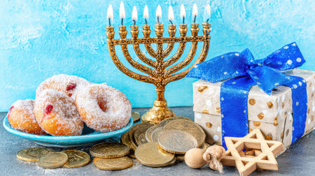 A vibrant Hanukkah celebration featuring a lit menorah, powdered sugar donuts, a gift box with a blue ribbon, chocolate coins, and wooden dreidels, all set against a blue background.の素材