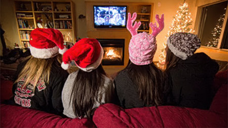 Four people wearing Santa hats and winter beanies watch a Christmas movie on TV by a cozy fireplace with a Christmas tree.の素材