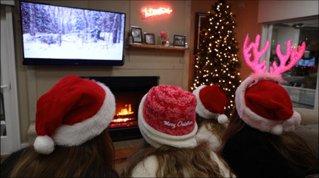 A family wearing Santa hats and reindeer antlers watches a Christmas movie on TV by a cozy fireplace with a decorated Christmas tree.の素材