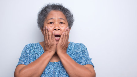 an older woman with gray hair is expressing shock with her hands on her face. she is wearing a blue shirt and has an open mouth in surpriseの素材