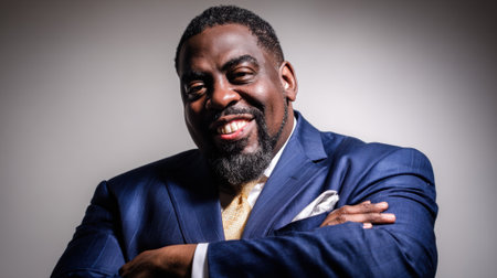 a confident african american man smiles in a blue suit with arms crossed against a light backdrop, exuding professionalism and success.の素材