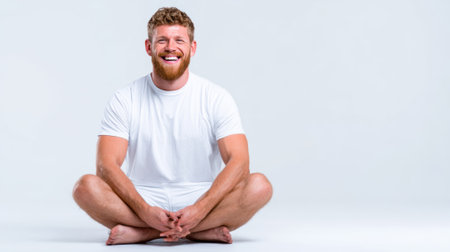 a cheerful man is sitting cross legged on the floor in a studio, wearing a white shirt, and smiling broadly at the camera with a light backgroundの素材