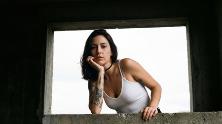 a portrait of a woman with tattoos in a white tank top, leaning on a concrete window frame, captured outdoors with a bright background and casual styleの素材