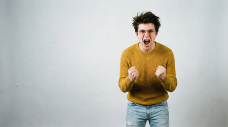 a man with glasses and a yellow sweater is yelling with his clenched fists. he is standing against a white background and appears to be angry.の素材