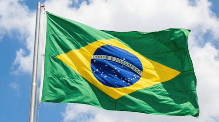 the Brazilian flag, with its vibrant green, yellow, and blue colors, waves gracefully against a backdrop of a clear blue sky dotted with fluffy cloudsの素材