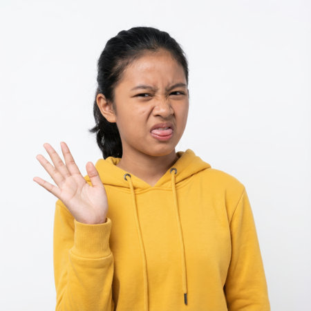 a young girl in a yellow hoodie expresses disgust with a grimace, tongue out, and a rejecting hand gestureの素材