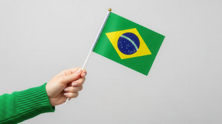 A person hand holding a small Brazilian flag against a plain, light gray background. The flag is green with a yellow diamond and a blue circle.の素材