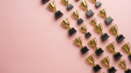 A top-down shot showcases numerous golden trophies meticulously arranged diagonally across a soft pink surface, symbolizing achievement and recognition.の素材