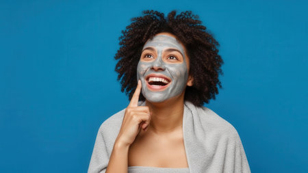 A joyful woman with curly hair applies a clay face mask, looking up with a smile, indicating a positive skincare experience.の素材