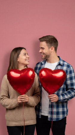 A happy couple stands close together, each holding a large red heart-shaped balloon and smiling at each other against a pink background.の素材