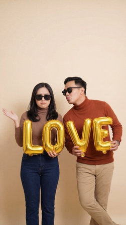 A couple poses playfully, holding gold balloons that spell out LOVE, while wearing sunglasses against a plain background.の素材