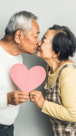 An elderly Asian couple shares a loving kiss while holding a pink heart, symbolizing their enduring affection.の素材